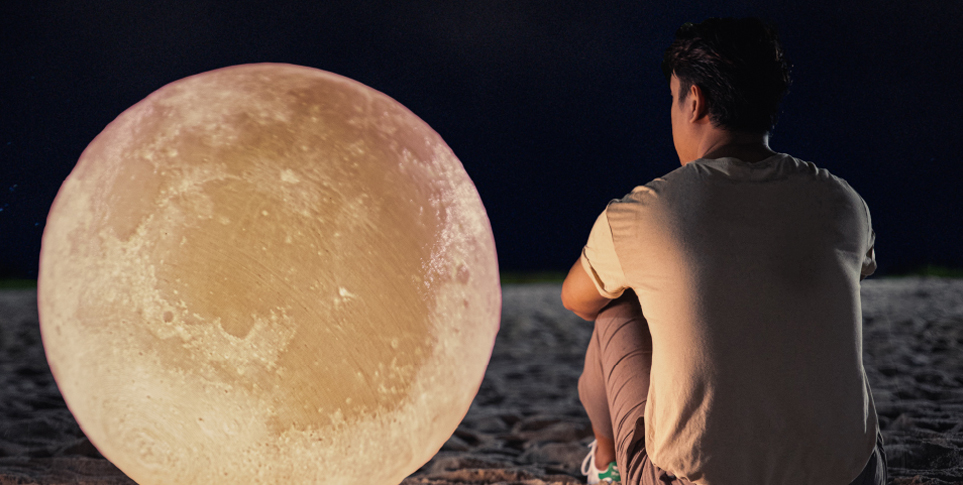 A man, Azim, is sitting on sand beside a large glowing moon, back-facing the camera. The sky around him is pitch black.