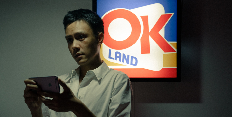 A young man in white collared shirt holds an iPhone but is looking ahead. Behind him is a lit sign that says "OK Land".