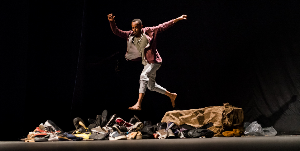 A young male refugee takes a leap over piles of shoes.