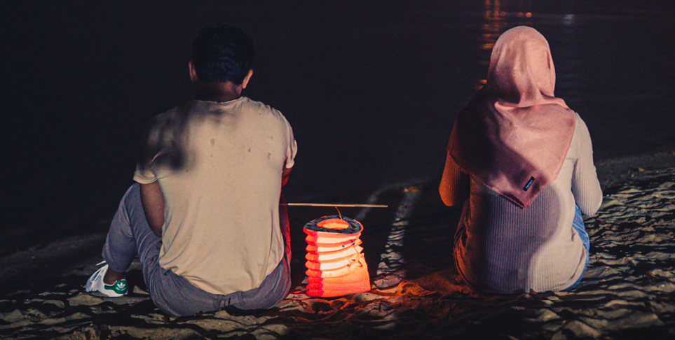 Azim and Mak sit on the sand with a lit Chinese lantern between them. They face the dark sea and ship lights.